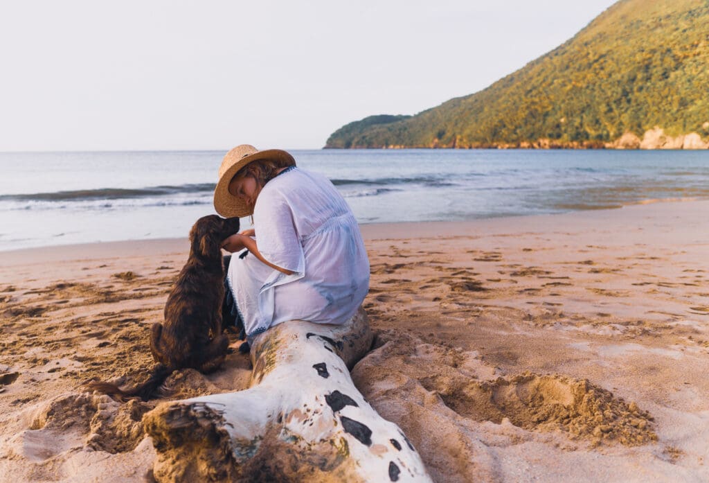 Blair sits on a log on a beach. A small brown dog sits next to her and she scratches under its chin.