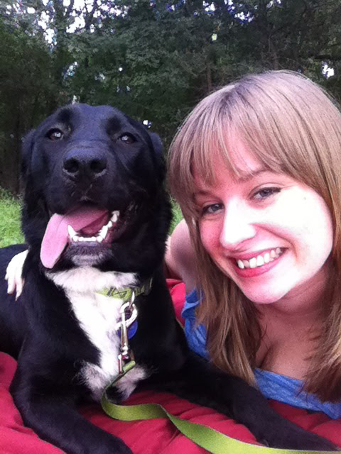 Blair, a young woman with dirty blonde hair, and Mia, a black dog with a white chest, pose with their heads close together looking at the camera.