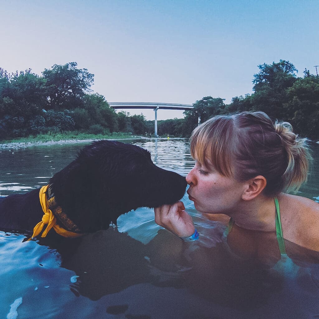 Blair, a young woman with blonde hair, and Mia, a black dog, are in a river. Blair's hand is under Mia's chin and Mia's nose touches Blair's lips.