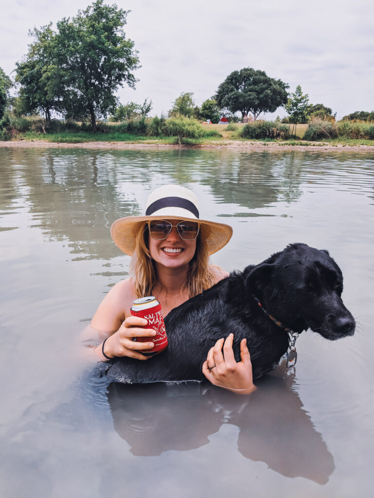 Blair wears large sunglasses, a hat, and holds a beer in the lake. Mia, a black dog, is on her lap.