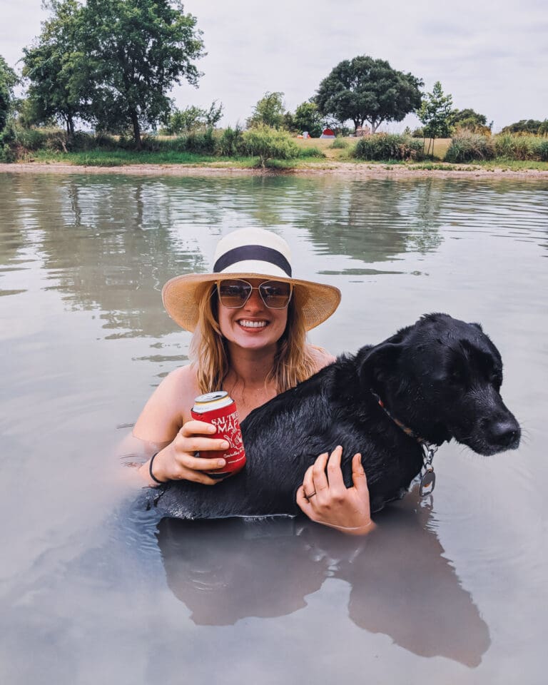 Blair wears a hat and holds a beer in the lake. Mia, a black dog, is on her lap.