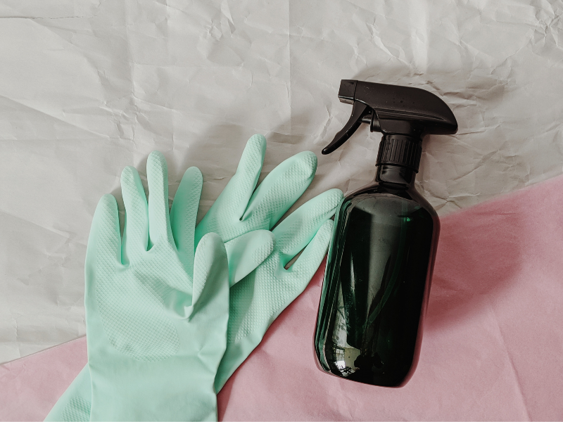 Light green gloves lay on wrinkled paper next to a brown glass spray bottle.
