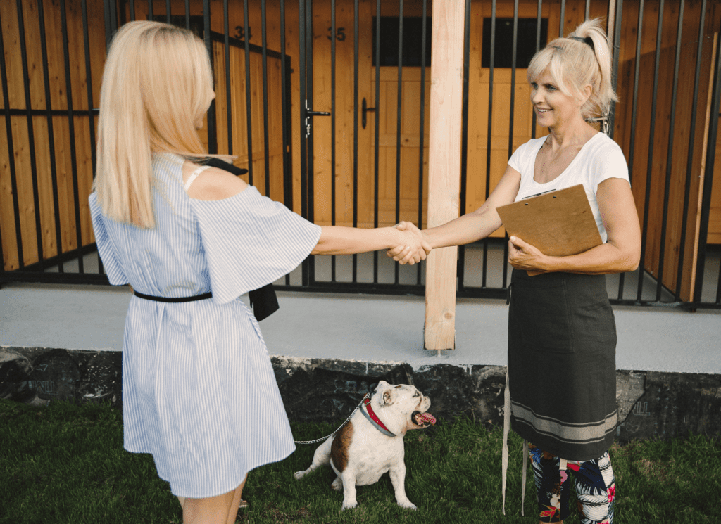 Two women shake hands at a dog boarding facility. One woman has a bulldog on a leash.