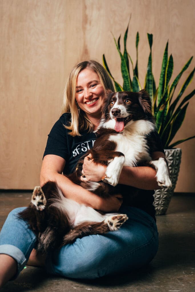 A woman sits on the ground holding her red and white Australian Shepherd, smiling. The dog has its mouth open and tongue hanging out.