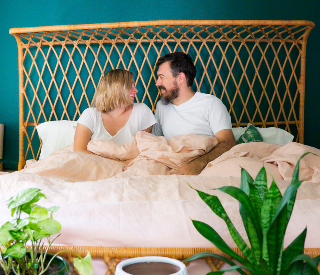 Husband and wife sit in bed laughing and smiling at each other.