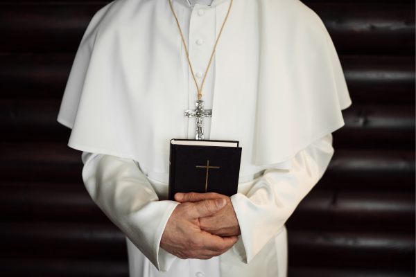 The Pop facing the camera wearing a gold chain rosary and holding a bible with both hands. His head is cropped out of th photo.