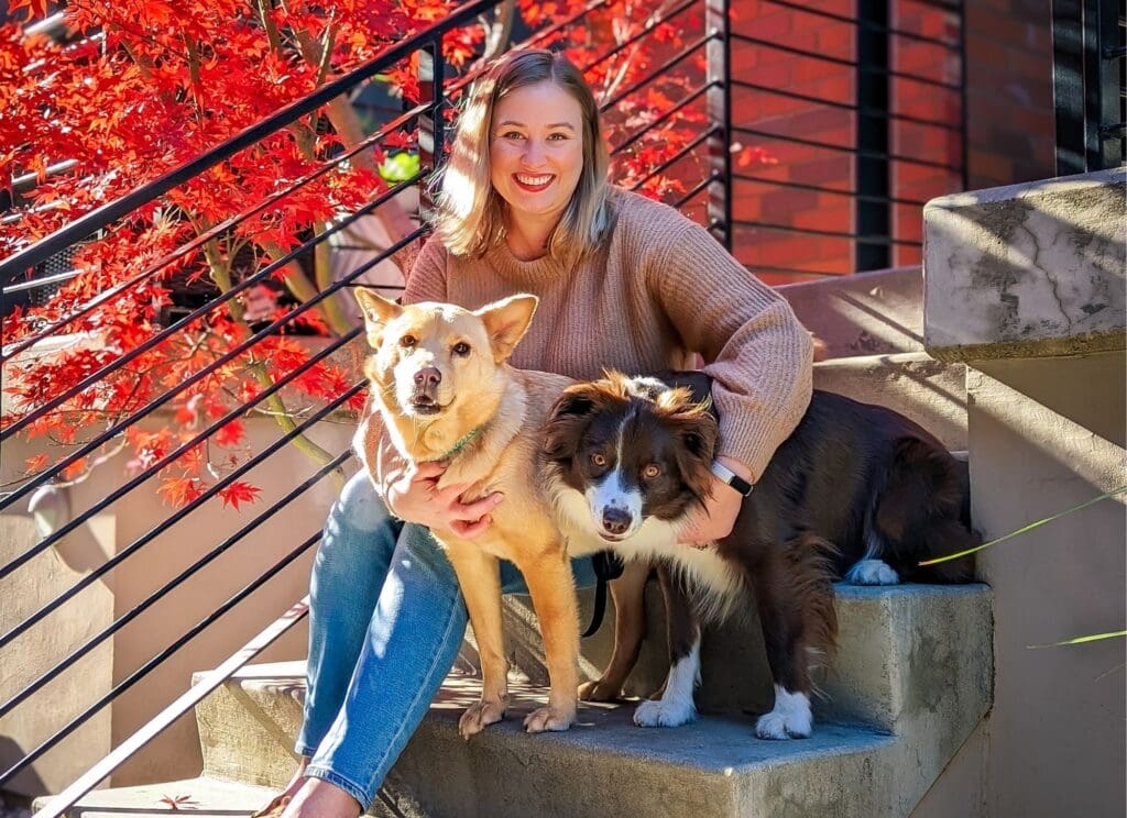 A blonde woman sits on concrete steps with two dogs.