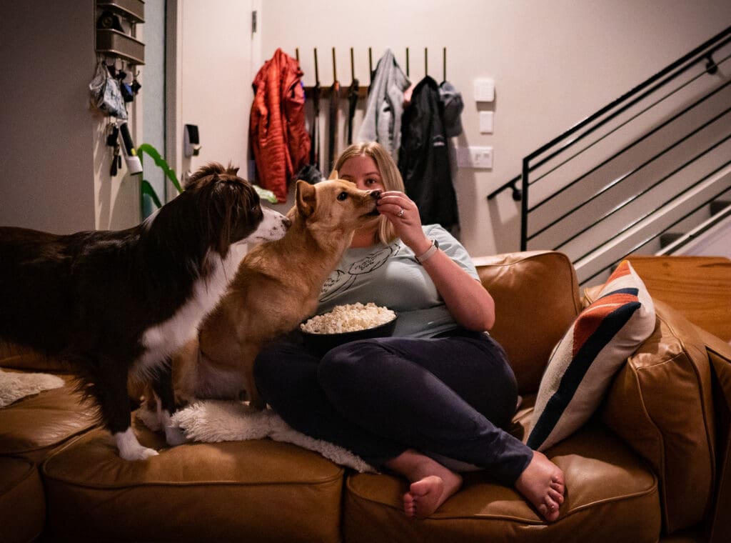 A girl sits on the couch with two dogs, feeding them popcorn.