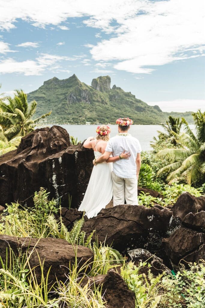 Blair and Brady stand among black volcanic rocks looking out towards a lush green mountain after their vow renewal ceremony.