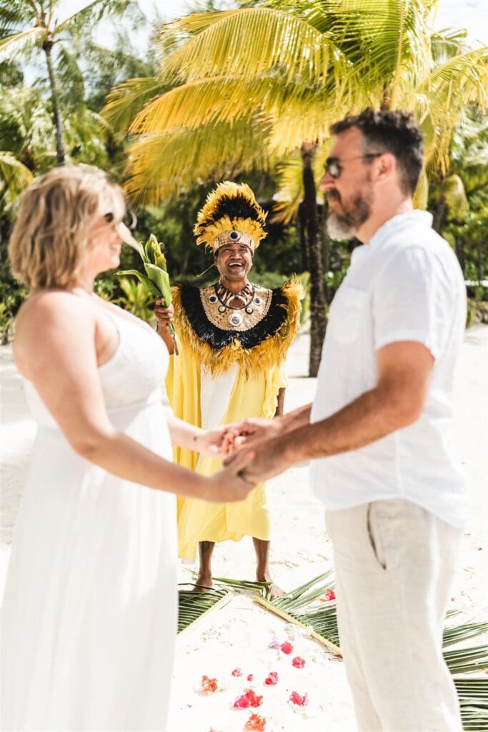 Blair and Brady are in the foreground, slightly out of focuse. They stand facing each other holding hands. Blair is in a white dress, Brady wears a white shirt and linen pants. A Polynesian Priest in traditional dress stands behind them, smiling and holding a handful of leaves as though blessing the ceremony.