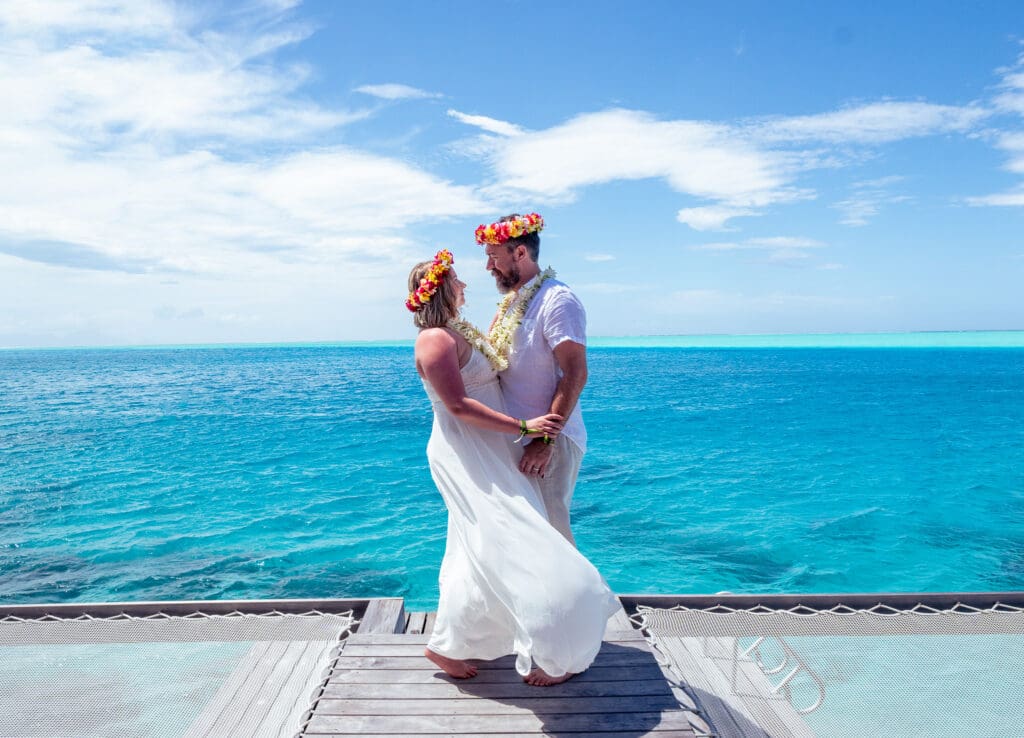 Blair and Brady, a heterosexual couple, stand on a grey dock overlooking turquoise waters. Both are dressed in white with bright colorful flower crowns, and they stare into each other's eyes.