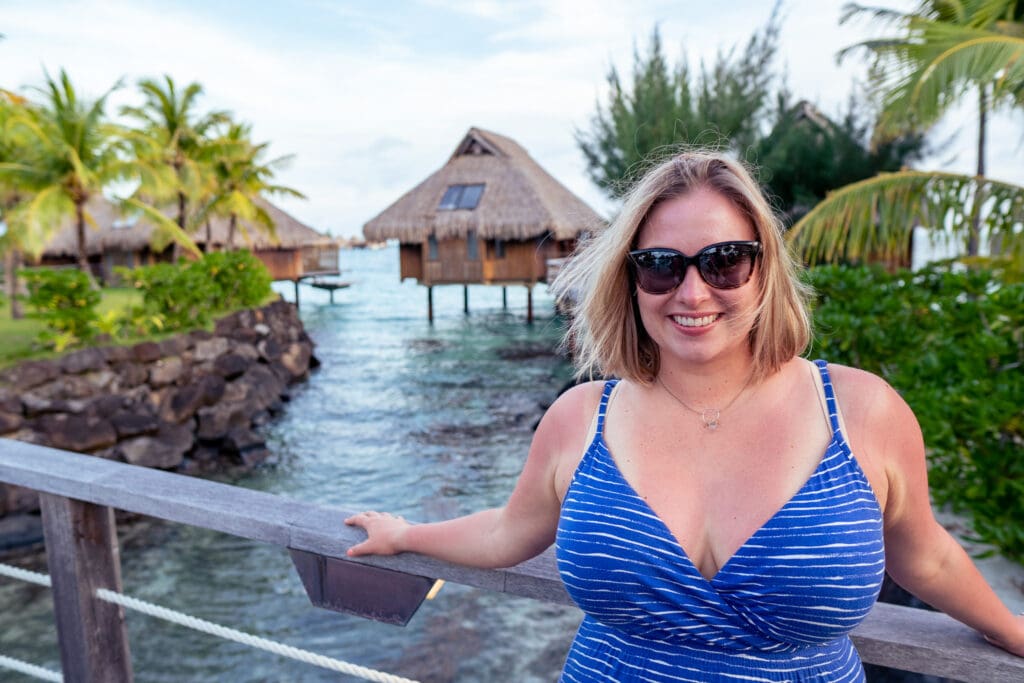 Blair, a white woman with shoulder length blonde hair, smiles at the camera. She is standing on a bridge over water with overwater bungalows and tropical foliage in the background. She wears  blue dress and brown Maui Jim sunglasses.