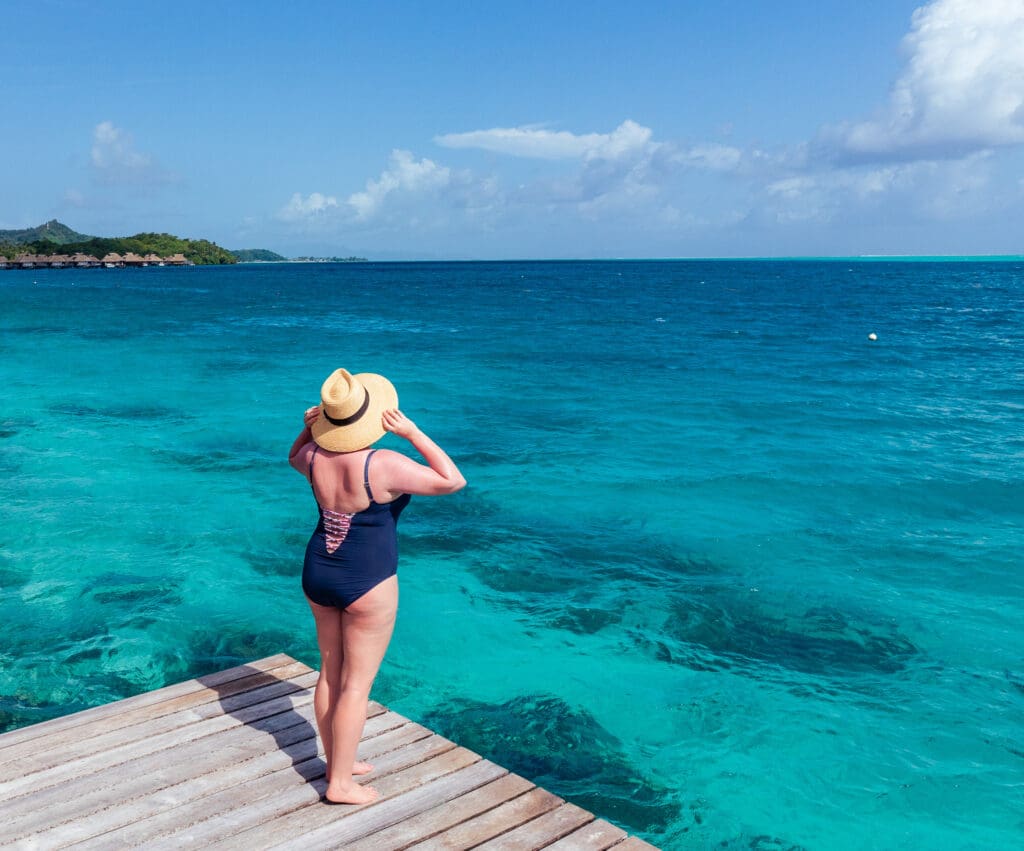 Blair, a white woman, stands on a wooden deck overlooking turquoise waters. She is wearing a navy one-pieec swimsuit with a colorful criss-cross back, as well as a broad-brimmed straw hat with a black band.