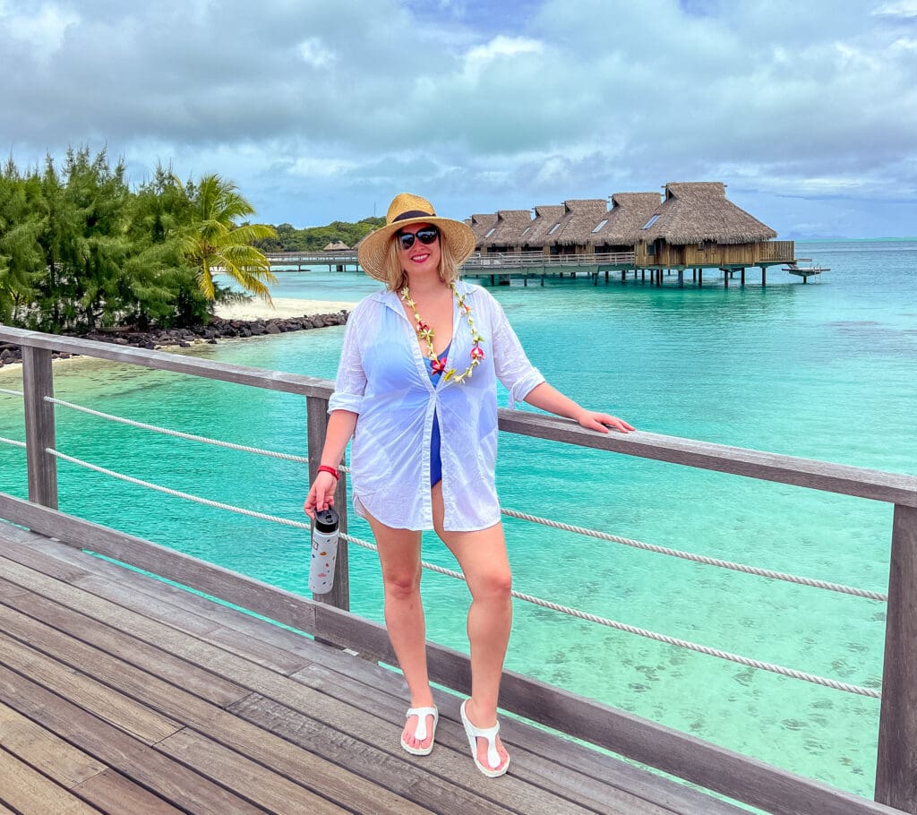 Blair stands on a bridge over turquoise water with overwater bungalowa in the background. She wears a straw sun hat, blue one-piece swimsuit, and a swimsuit cover up that looks like an oversized button down shirt.
