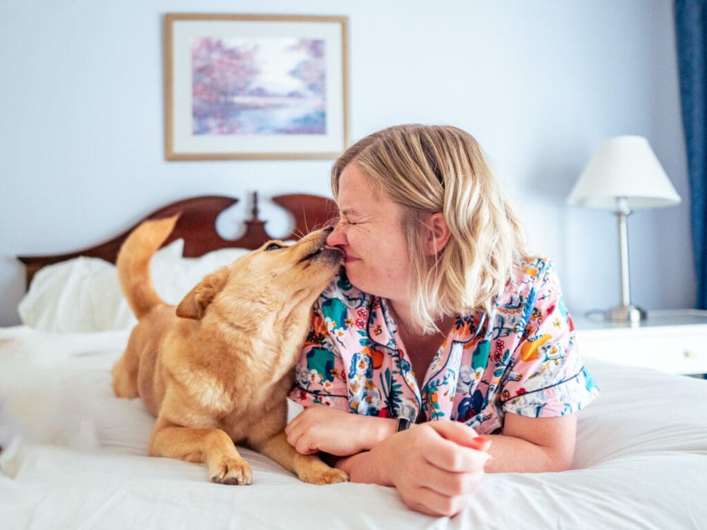 Blair lays on her stomach on a bed in a dog friendly hotel room at the Sands Inn & Suites in San Luis Obispo. Her dog, a yellow mutt named June, lays next to her and is kissing her face.