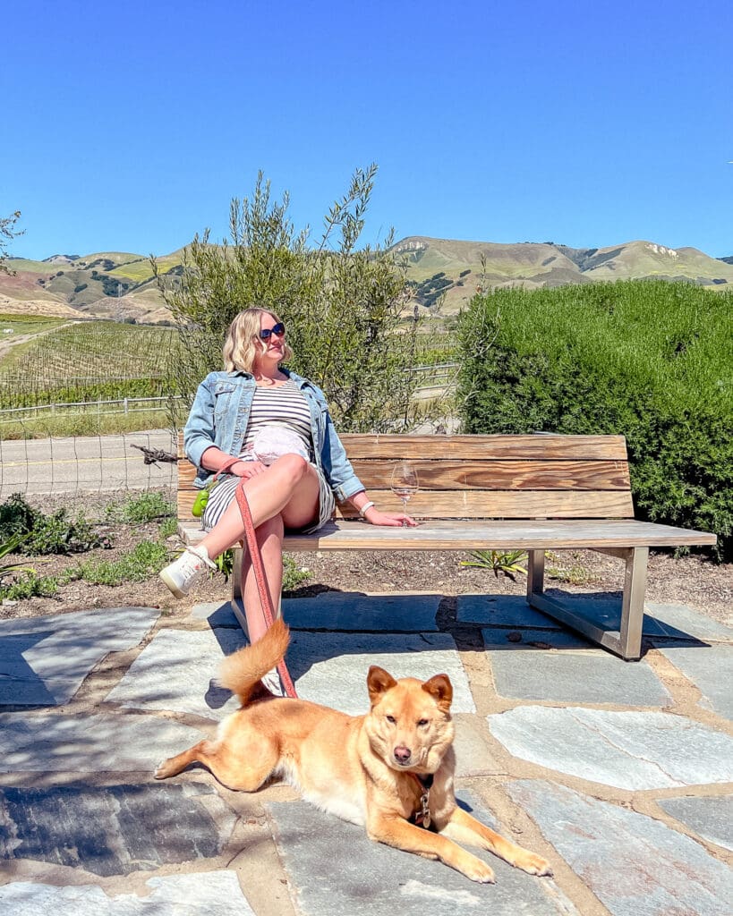 Blair sits on a bench at Chamisal Vineyard in San Luis Obispo with a glass of wine. June, a yellow mutt, lays on the ground at her feet. Behind them are rolling hills.